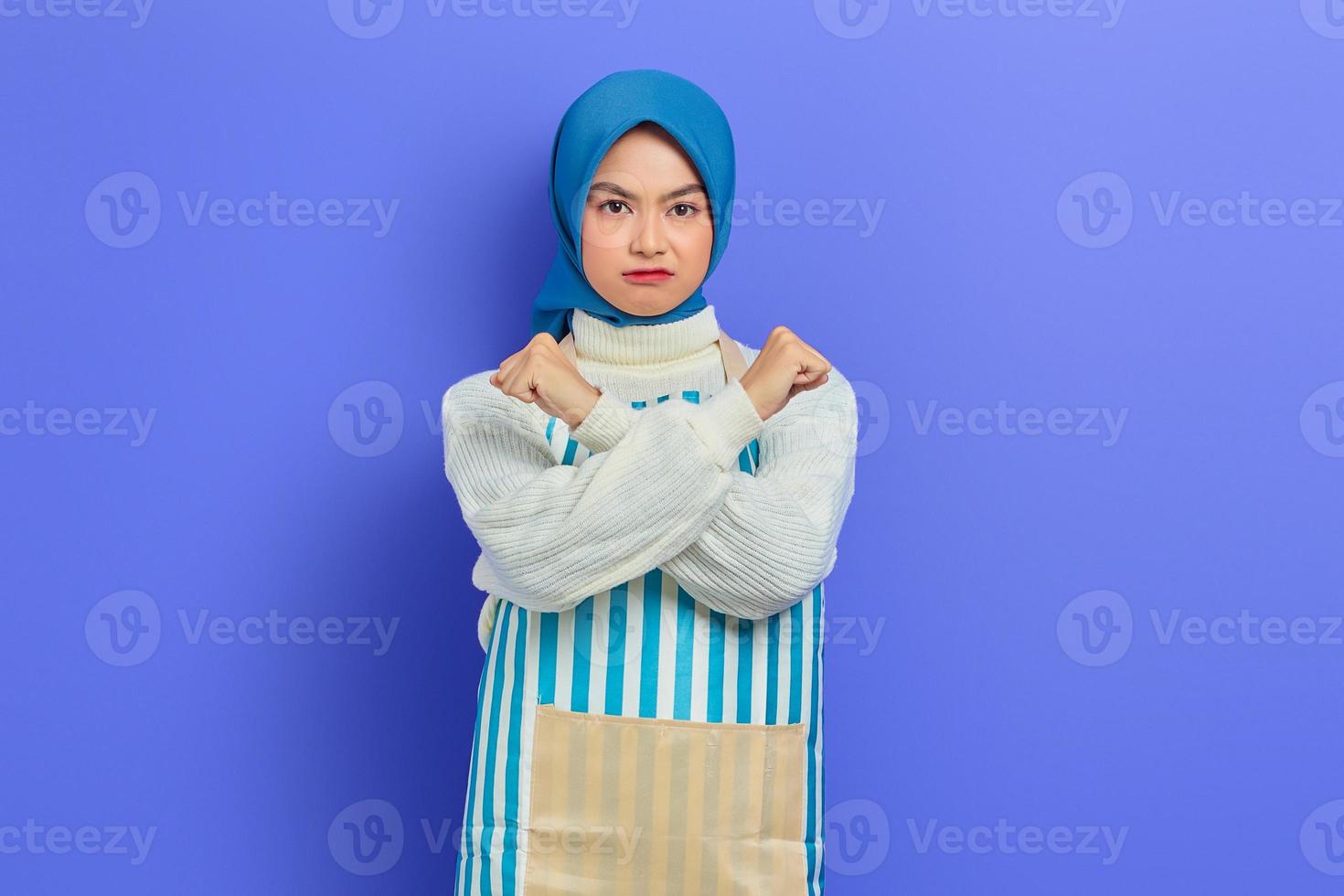 Ritratto di giovane casalinga arrabbiata in hijab e grembiule a righe che fa un gesto di arresto con le mani incrociate isolate su sfondo viola. concetto di stile di vita musulmano casalinga di persone foto