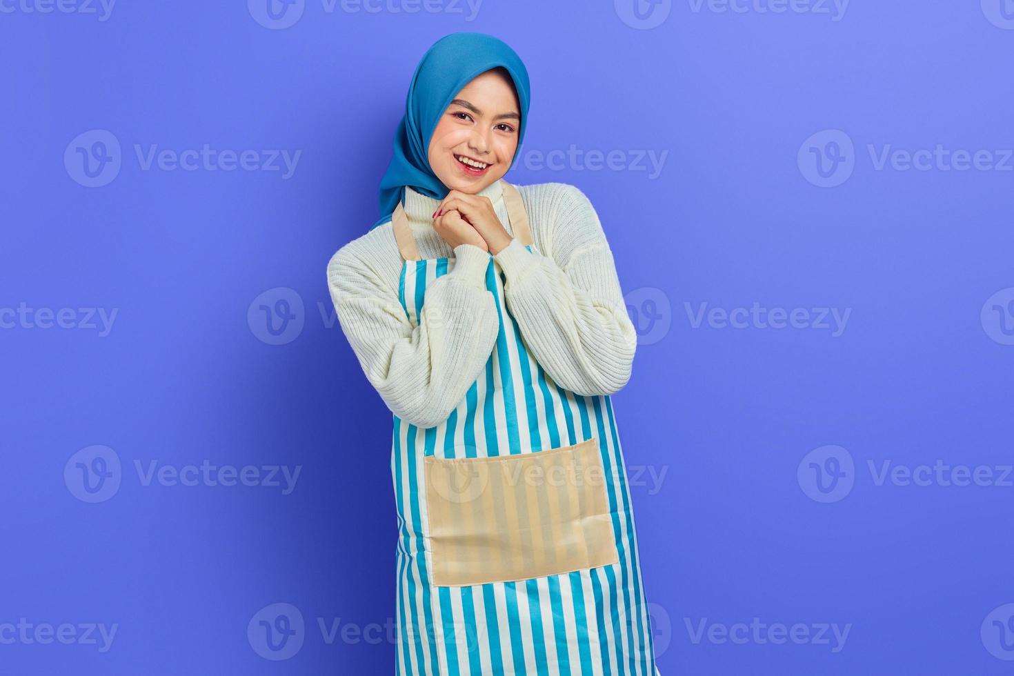 ritratto di donna casalinga sorridente in hijab e grembiule sfregandosi le mani e guardando la fotocamera isolata su sfondo viola. concetto di stile di vita musulmano casalinga di persone foto