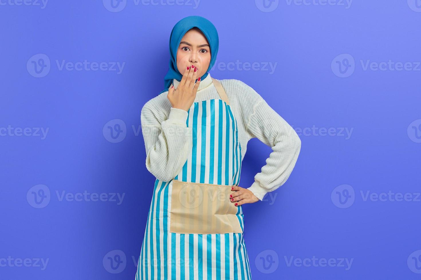 ritratto di giovane casalinga scioccata in hijab e grembiule che copre la bocca con le mani isolate su sfondo viola. concetto di stile di vita musulmano casalinga di persone foto