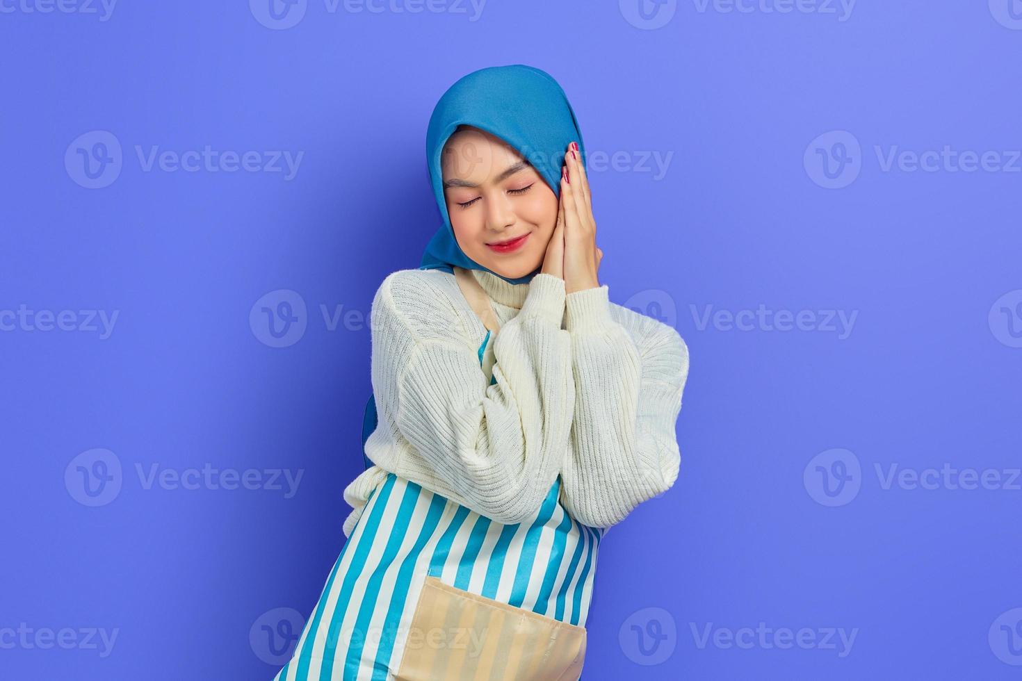 Ritratto di giovane donna musulmana asiatica sorridente in hijab e grembiule a righe, che dorme con le mani giunte sotto le guance e gli occhi chiusi isolati su sfondo viola foto
