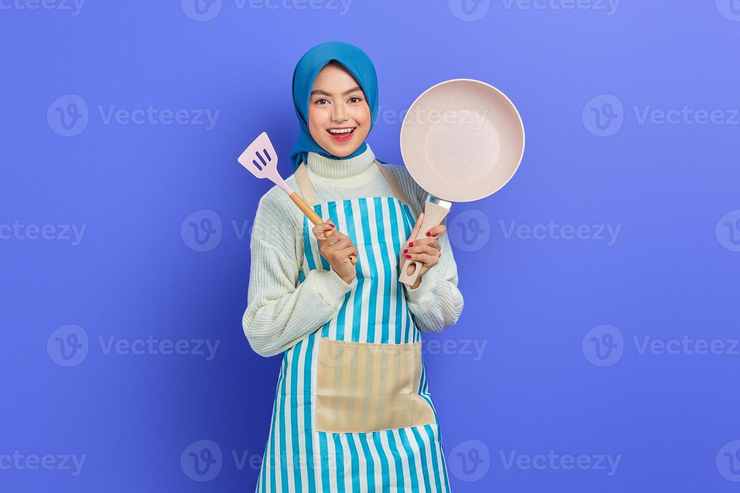 sorridente giovane donna asiatica casalinga che indossa un grembiule tenendo la spatola e la padella per friggere mentre fa i lavori domestici isolati su sfondo viola. concetto di pulizia foto