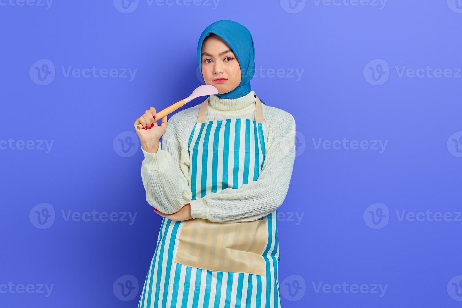 bella donna asiatica pensierosa in maglione bianco ricoperta di grembiule e hijab che tiene un cucchiaio, guardando la fotocamera facendo i lavori domestici isolati su sfondo viola. concetto di pulizia foto