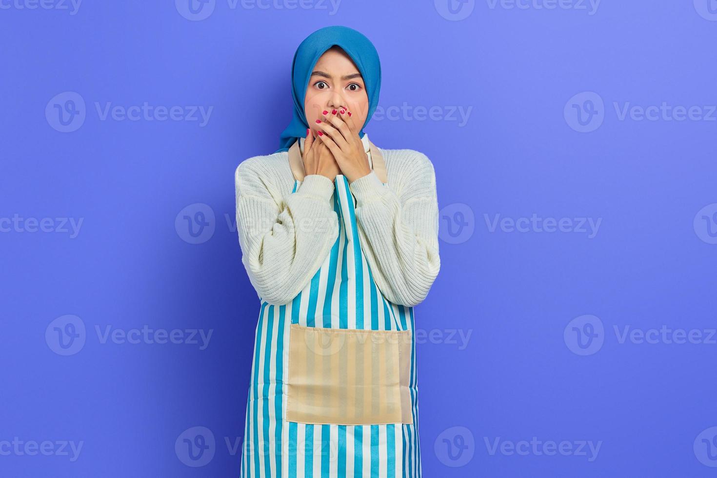 ritratto di giovane donna casalinga spaventata in hijab e grembiule guardando la fotocamera mentre si copre la bocca con le mani isolate su sfondo viola. concetto di stile di vita musulmano casalinga di persone foto