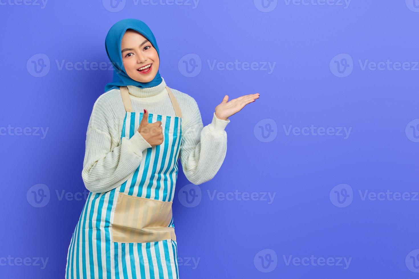 sorridente giovane donna musulmana asiatica in hijab e grembiule a righe, indicando lo spazio della copia con le mani, mostrando il gesto del pollice in su isolato su sfondo viola. concetto di stile di vita musulmano casalinga di persone foto