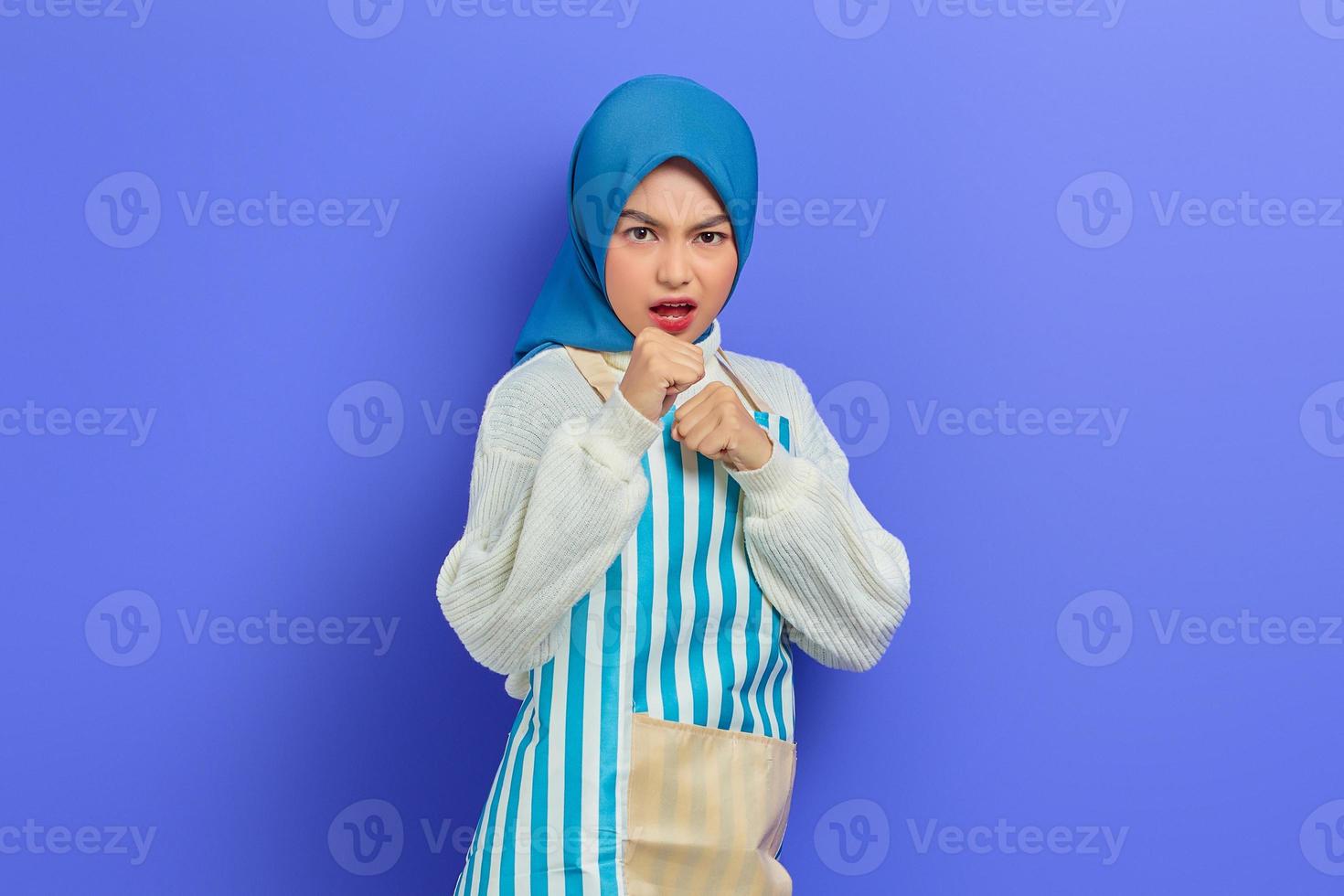 ritratto di giovane donna musulmana asiatica dispiaciuta in hijab e grembiule a righe in piedi pugni serrati boxe e fotocamera guardando isolata su sfondo viola. concetto di stile di vita musulmano casalinga di persone foto
