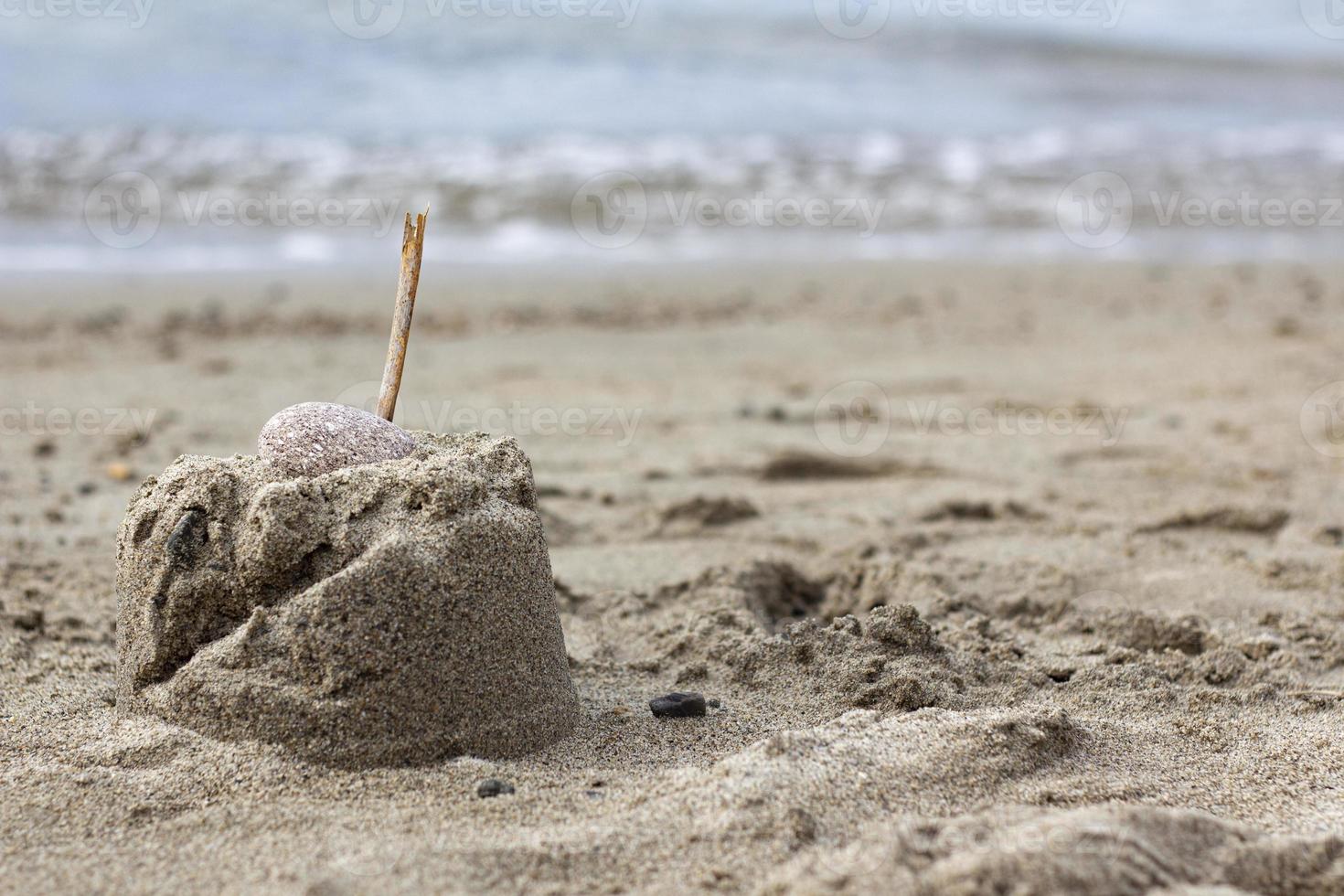 montagna di sabbia con un bastone di legno sulla spiaggia. castello di sabbia con un sasso sulla spiaggia. primo piano di piccoli castelli di sabbia con un bastone sormontato da un cielo blu foto