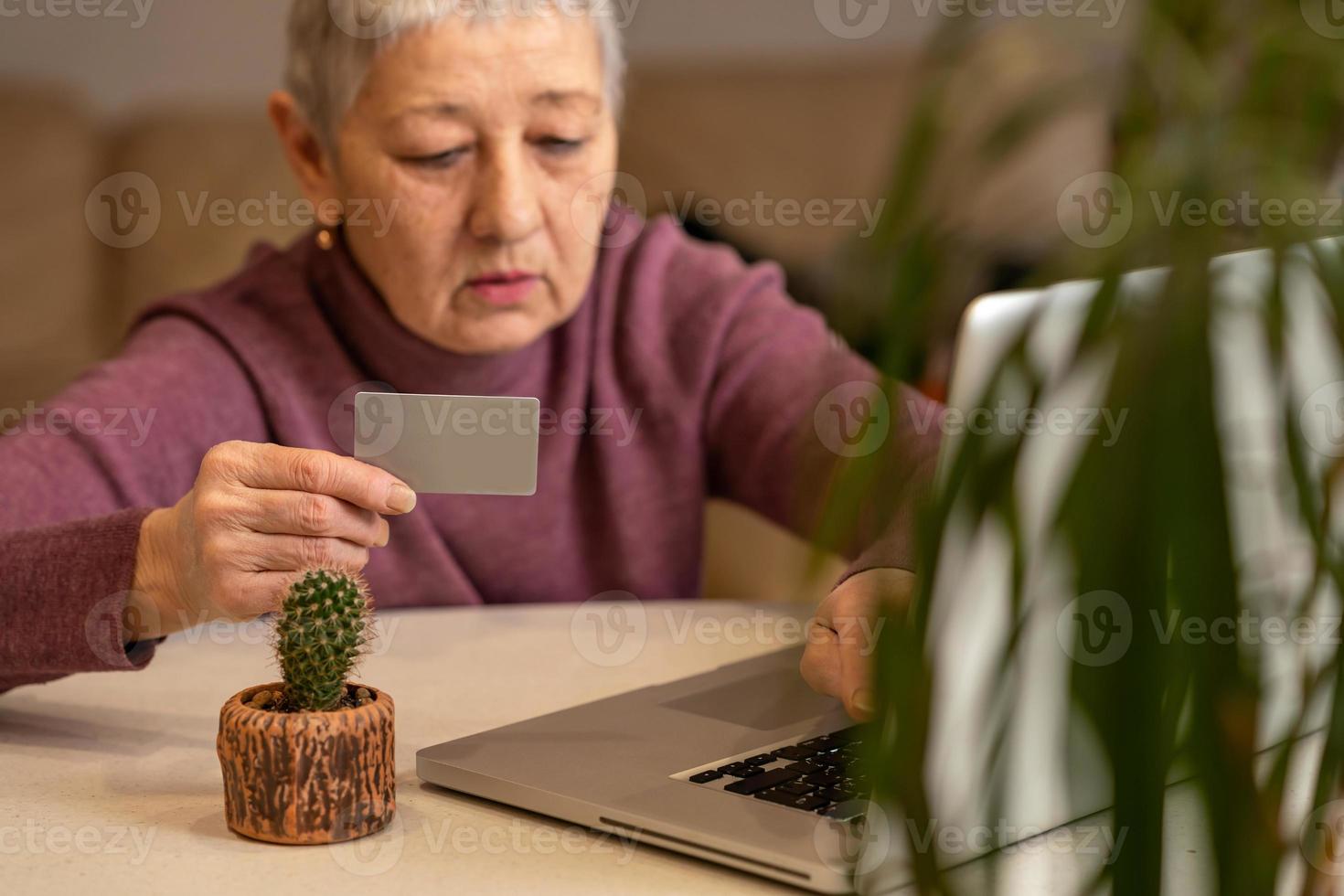 una donna anziana con i capelli grigi si siede davanti a un laptop con una carta di credito in mano, fa acquisti online. concetto di acquisto online foto