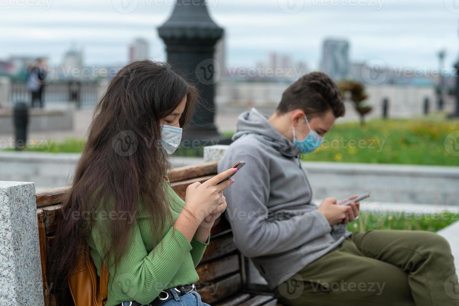 distanza sociale, un ragazzo e una ragazza sono seduti su una panchina a distanza l'uno dall'altro con la mascherina sul viso e con i telefoni in mano foto