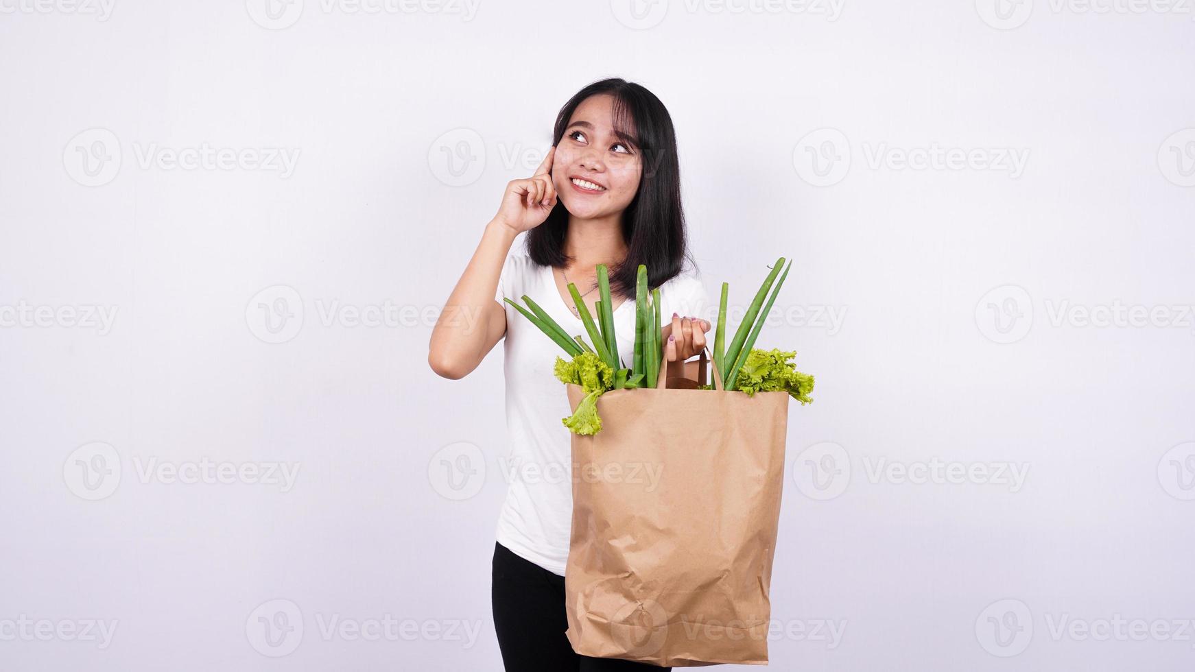 donna asiatica che pensa con il sacchetto di carta delle verdure fresche con fondo bianco isolato foto