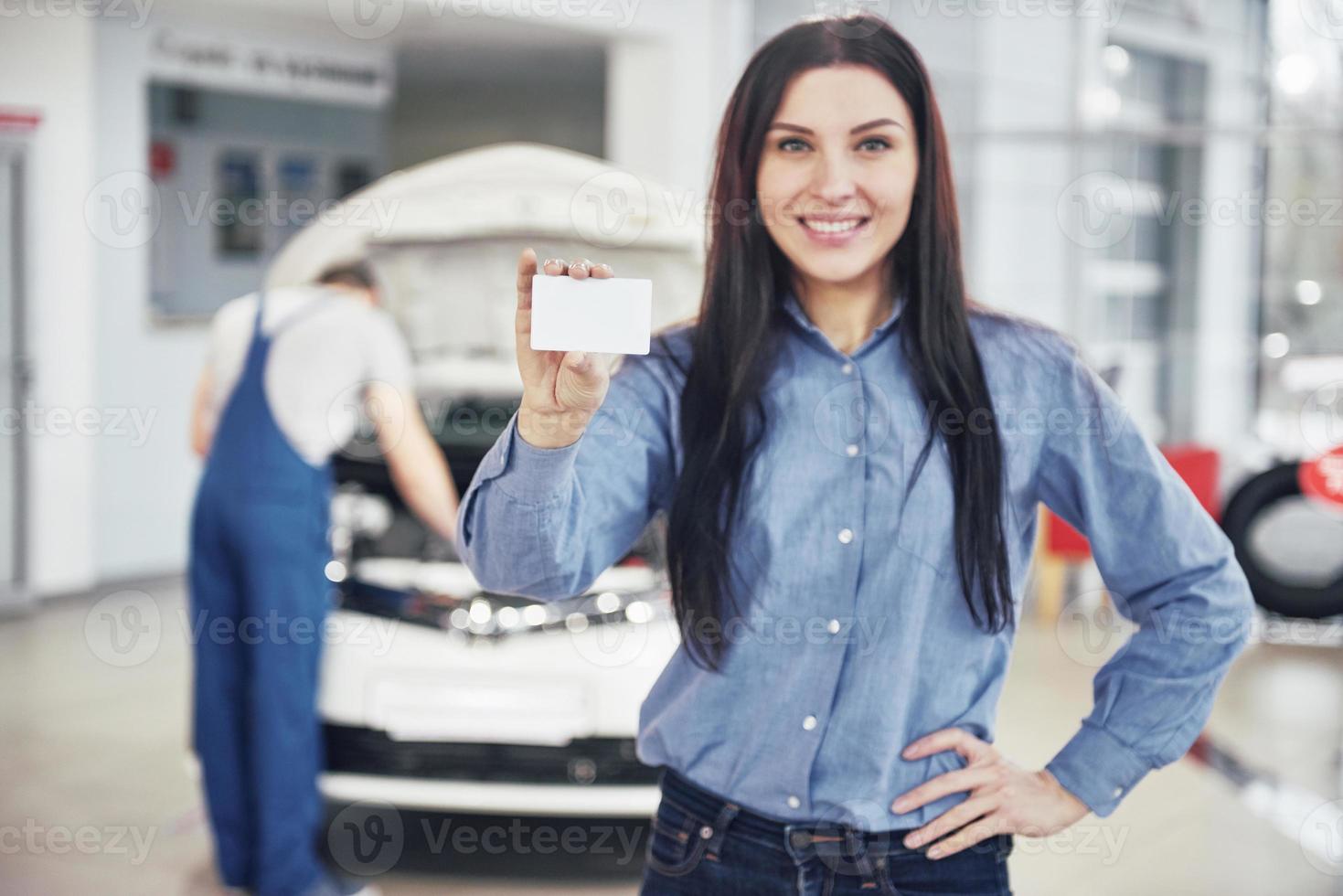 una bella donna conserva un biglietto da visita del centro servizi auto. il meccanico ispeziona l'auto sotto il cofano sullo sfondo foto