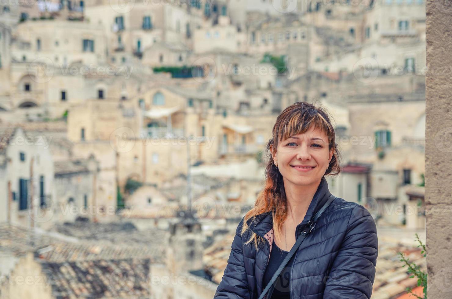 giovane bella donna viaggiatore con giacca nera che guarda l'obbiettivo, sorride, posa e mantiene la spighetta nei sassi di matera foto