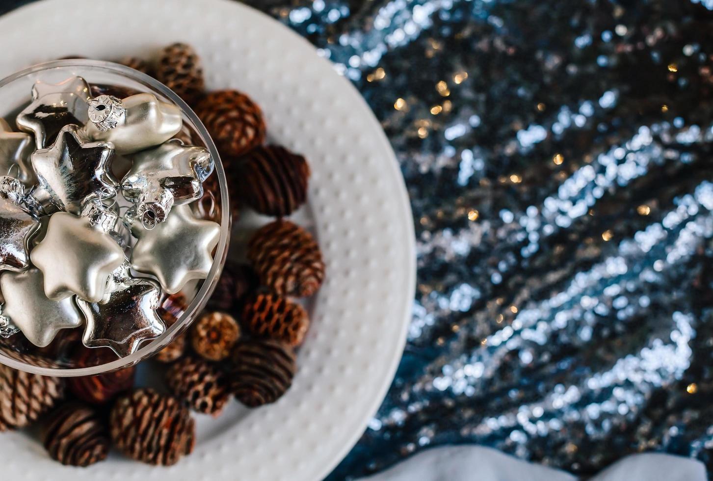 stelle d'argento di vetro in un vaso di vetro. giocattoli dell'albero di natale in un bicchiere su uno sfondo di tessuto lucido e pigne. copia spazio. foto