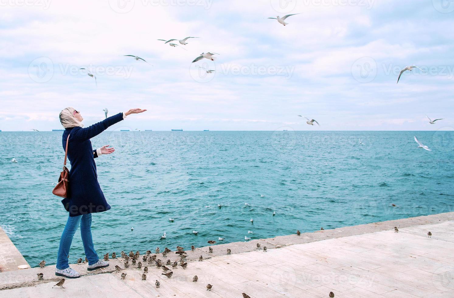 donna bionda che alimenta i gabbiani in una nuvolosa giornata autunnale sulla costa del mare foto