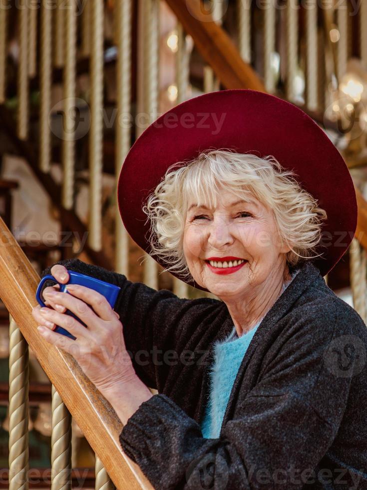 donna anziana alla moda in cappotto grigio scuro, cappello e con i capelli grigi in piedi accanto alla giostra sorridendo, bevendo caffè e godendosi la vita. viaggio, divertimento, pensione, felicità, concetto stagionale foto