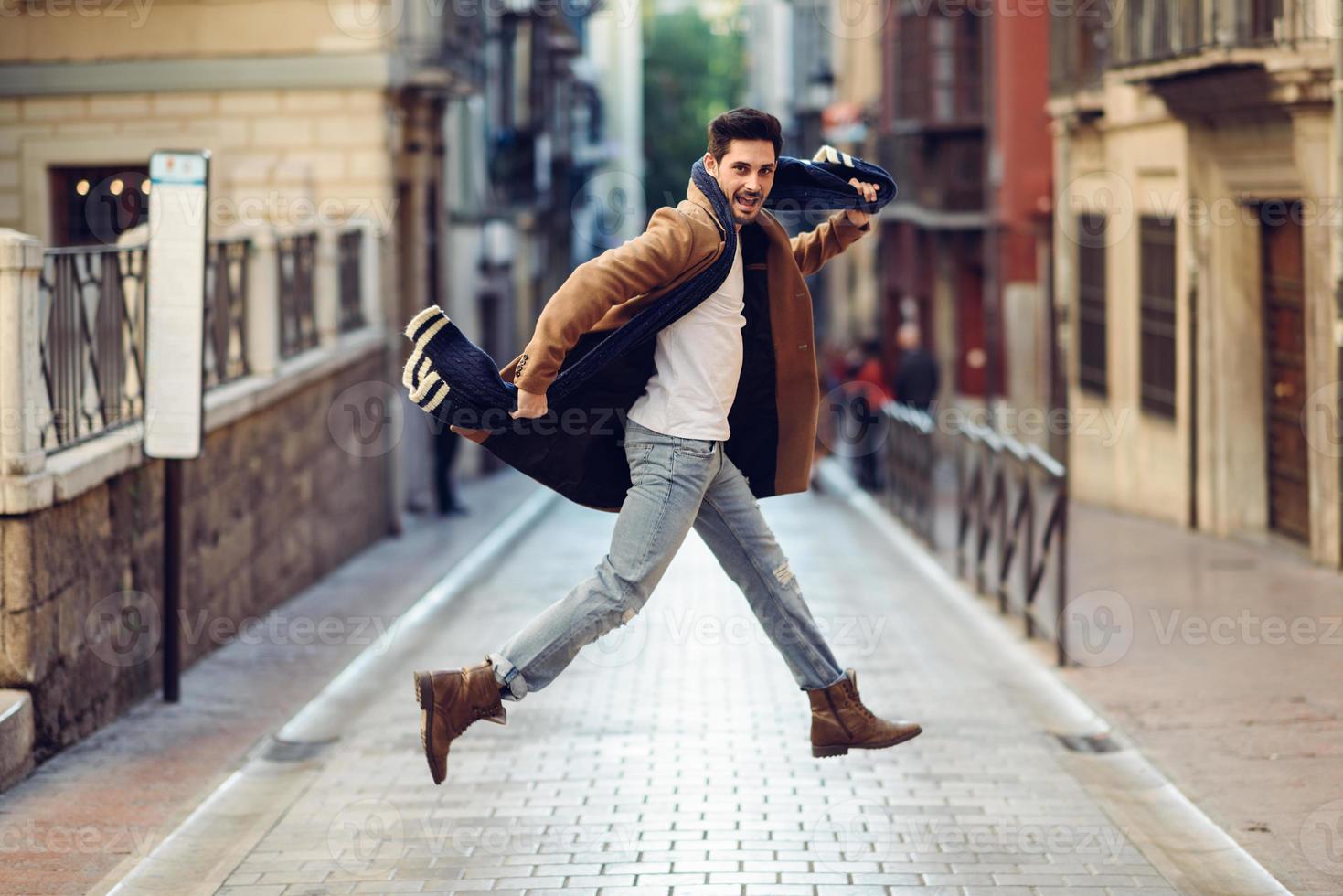 giovane uomo felice che salta indossando abiti invernali in background urbano foto