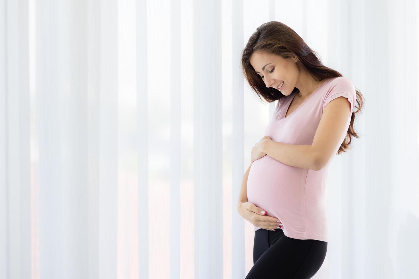 felice giovane e bella donna incinta tocco tenero pancia con amore, cura e felicità. nuova madre in piedi vicino al sipario con speranza, sognando come maternità. profonda emozione del concetto di felicità foto