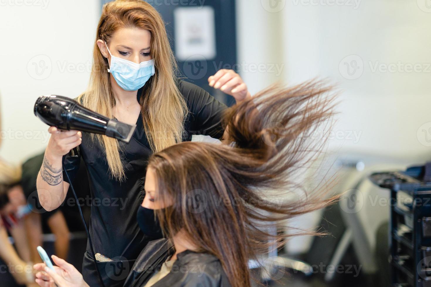 parrucchiere che asciuga i capelli della sua cliente con un asciugacapelli indossando maschere protettive in un centro estetico. foto