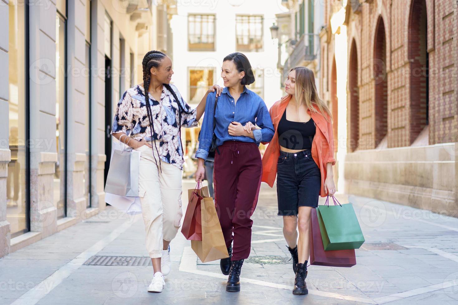 maniaci dello shopping femminili multirazziali alla moda che camminano insieme per strada con sacchetti di carta foto