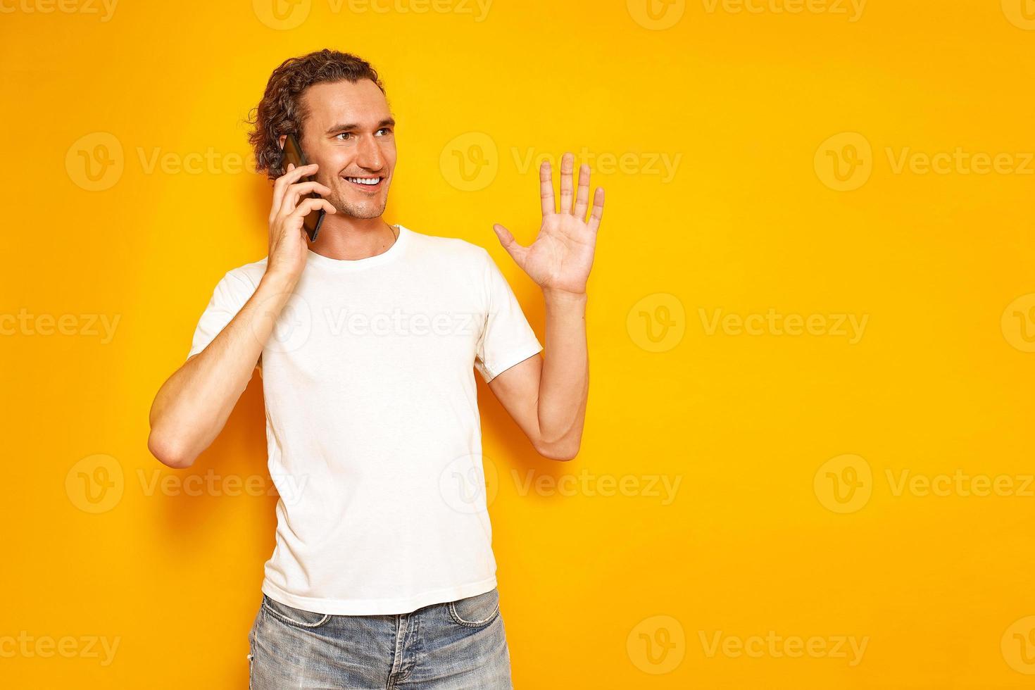 l'uomo sorridente sta parlando al telefono gesticolando con la mano alzata, godendosi la comunicazione, vestito con abiti casual. isolato su sfondo giallo studio. concetto - persone, tecnologia, comunicazione foto