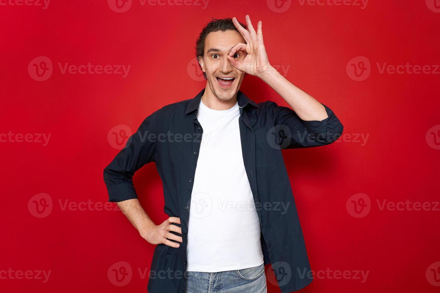 l'uomo eccitato con la bocca aperta tiene la mano sotto forma di un segno ok vicino all'occhio e guarda attraverso le dita. vestito con abiti casual isolati su sfondo rosso per studio. concetto - persone, approvazione, vendita foto