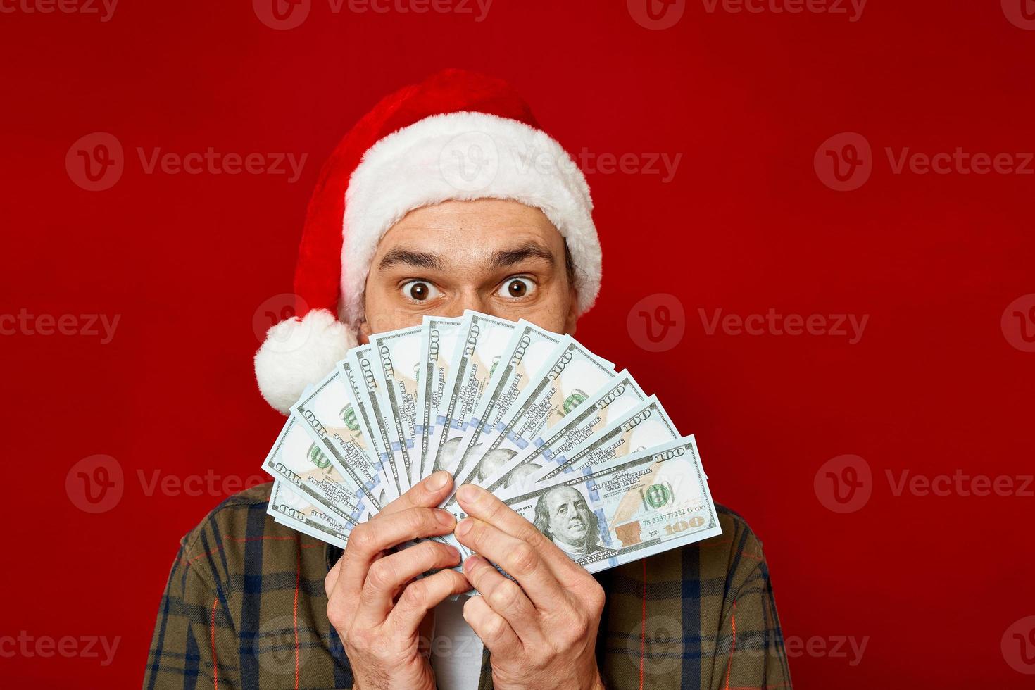 grande ritratto in studio di giovane uomo con cappello di Natale con mazzetta di denaro nelle sue mani che tiene un fan di dollari davanti al suo viso in stato di shock per la vittoria alla lotteria di successo. concetto vincente, cash back, ricchezza foto