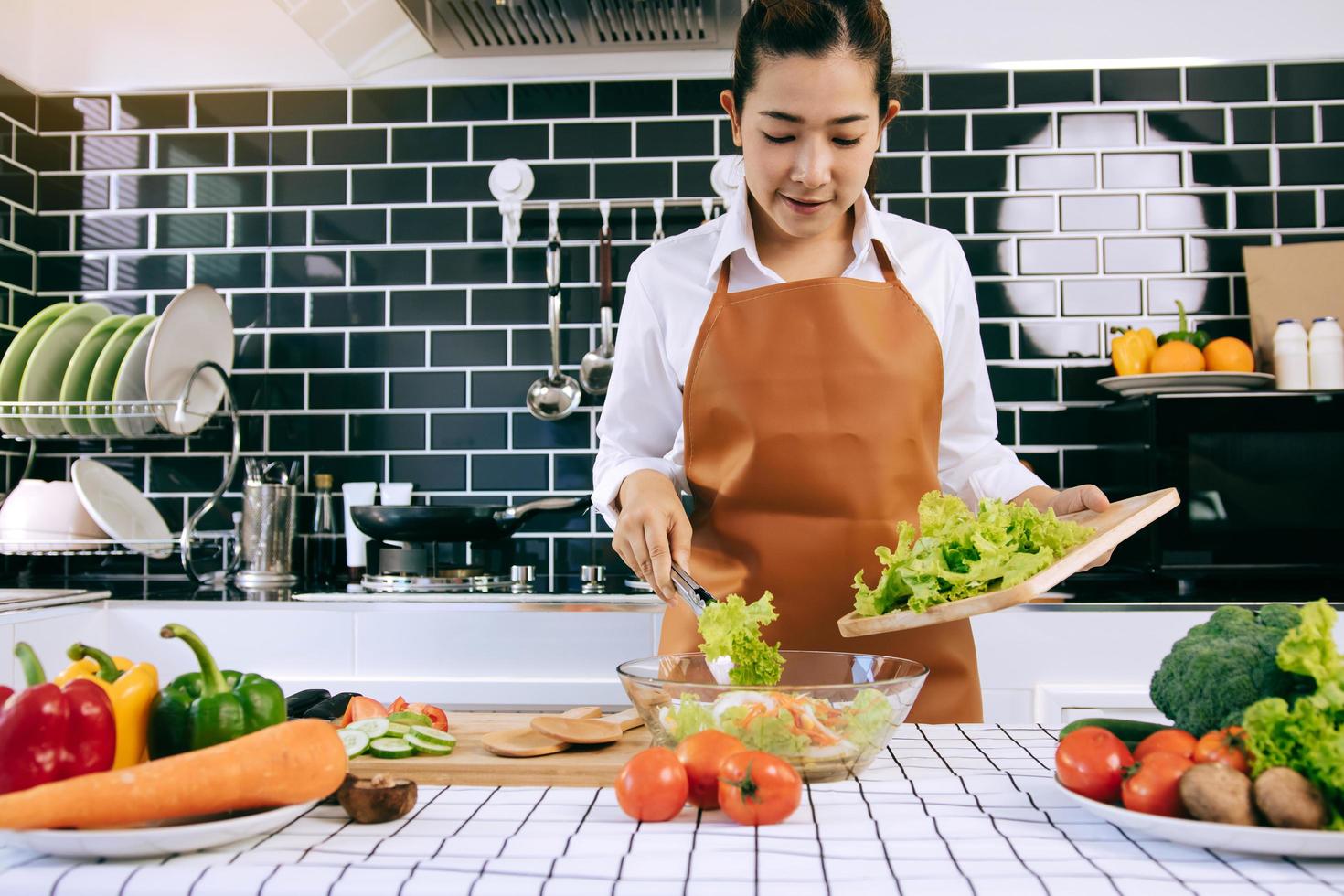 la casalinga asiatica sta usando le pinze per portare l'insalata sul tagliere di legno sulla tazza di insalata nella stanza della cucina. foto