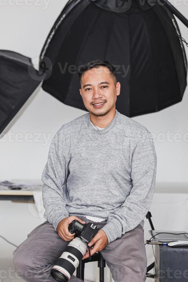 fotografo asiatico sorridente seduto e tenendo la fotocamera in studio foto