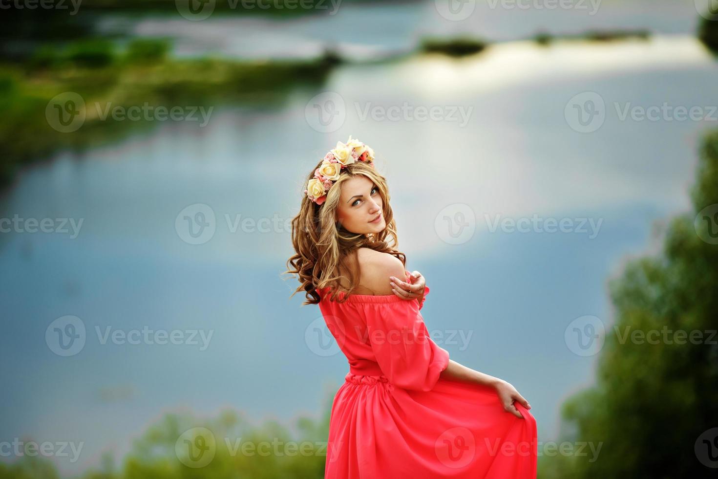 bella giovane donna bionda con una corona di fiori in testa. ragazza di bellezza con l'acconciatura di fiori in un abito lungo rosso vicino al lago. foto di moda, copia spazio