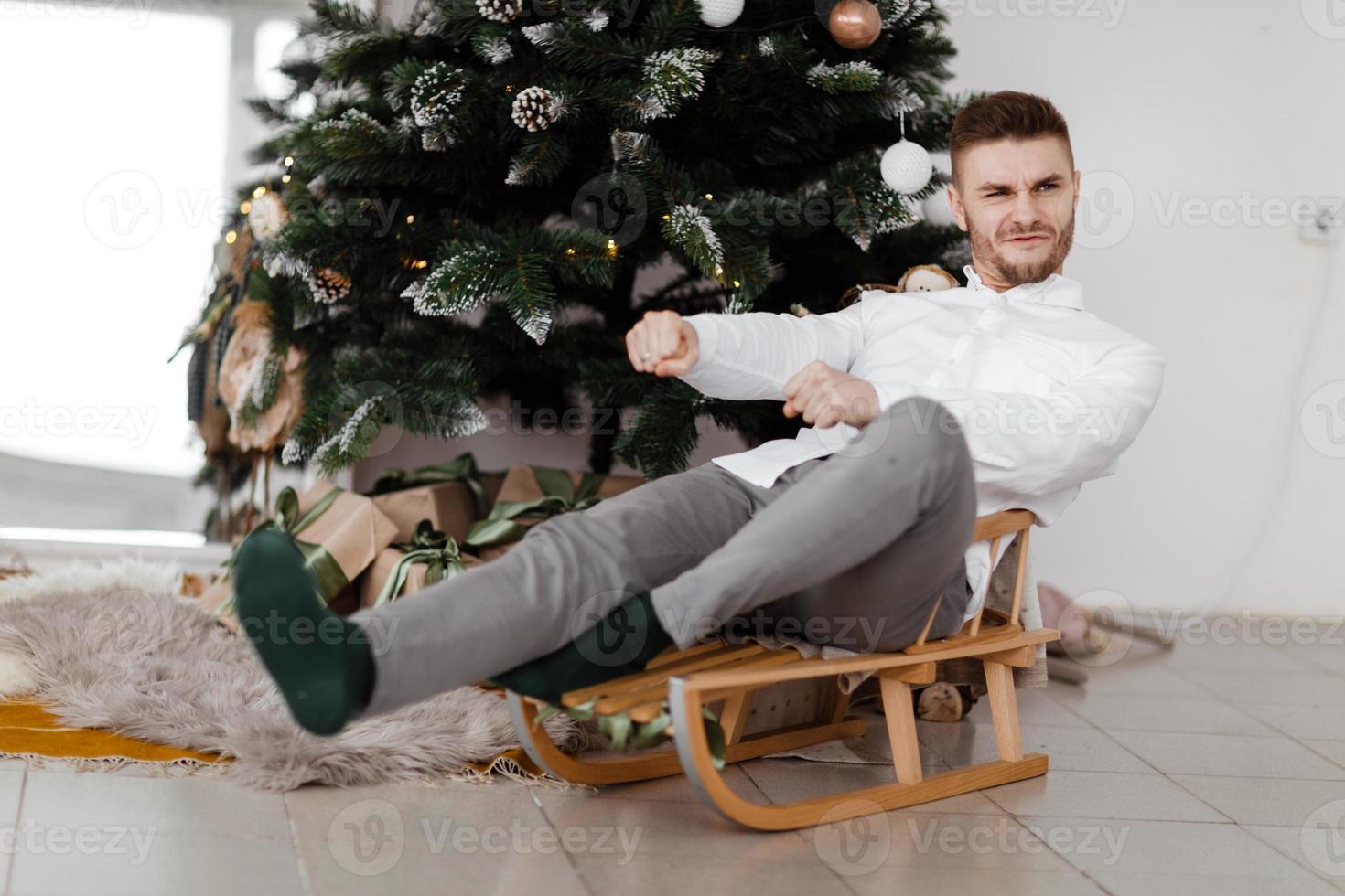 uomo allegro seduto sulla slitta a casa vicino all'albero di natale. uomo su una slitta. buon umore natalizio. concetto di famiglia e vacanza. foto