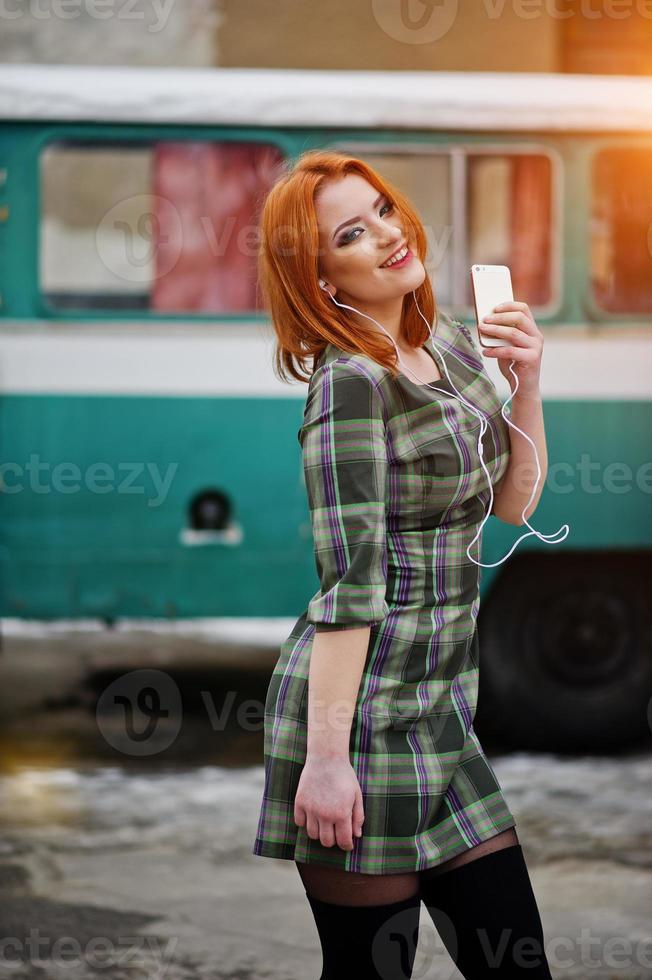 giovane ragazza dai capelli rossi signora con cellulare e cuffie, indossando su sfondo vestito controllato vecchio bus minivan turchese vintage. foto