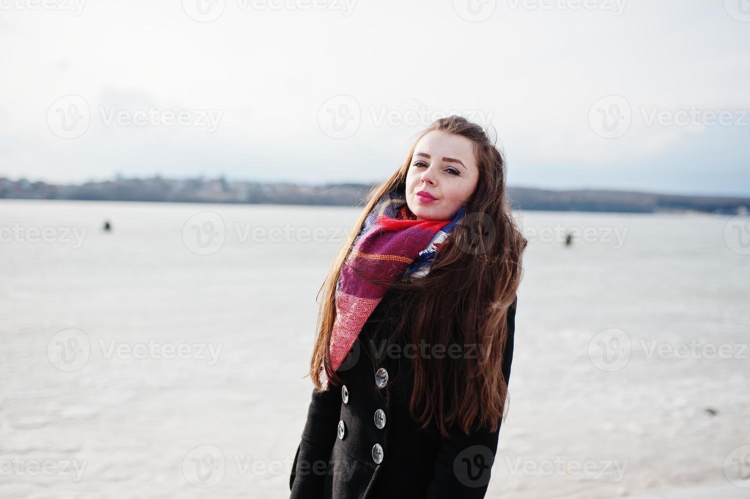 ragazza casual al cappotto nero, sciarpa e cappello contro il fiume ghiacciato in un clima invernale soleggiato. foto