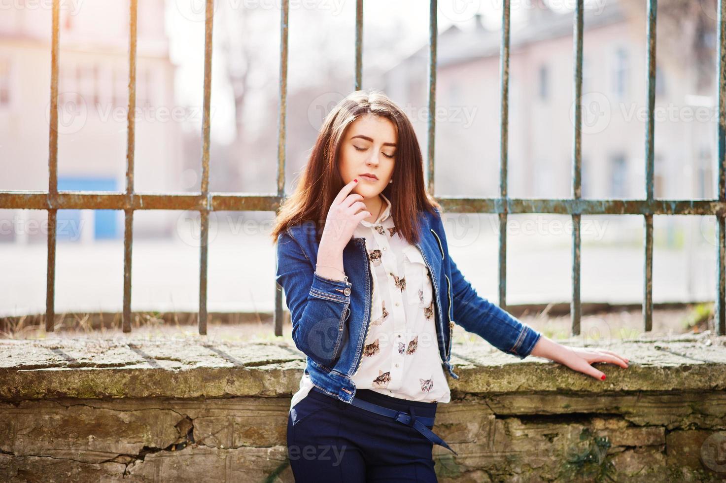giovane ragazza bruna alla moda su camicia, pantaloni, giacca di jeans posata sullo sfondo del recinto di ferro. concetto di modello di moda di strada. foto