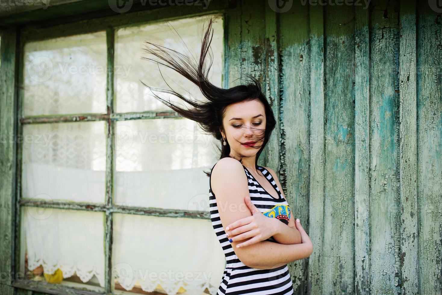 ragazza modello bruna con i capelli in aria che indossa un vestito con sfondo a strisce cian casa in legno con vecchie finestre. foto