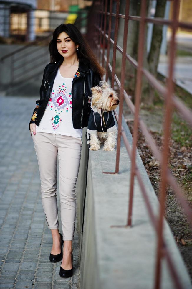 ragazza gitana bruna con cane yorkshire terrier poste contro ringhiere in acciaio. indossa il modello su giacca di pelle e t-shirt con ornamento, pantaloni e scarpe con i tacchi alti. foto