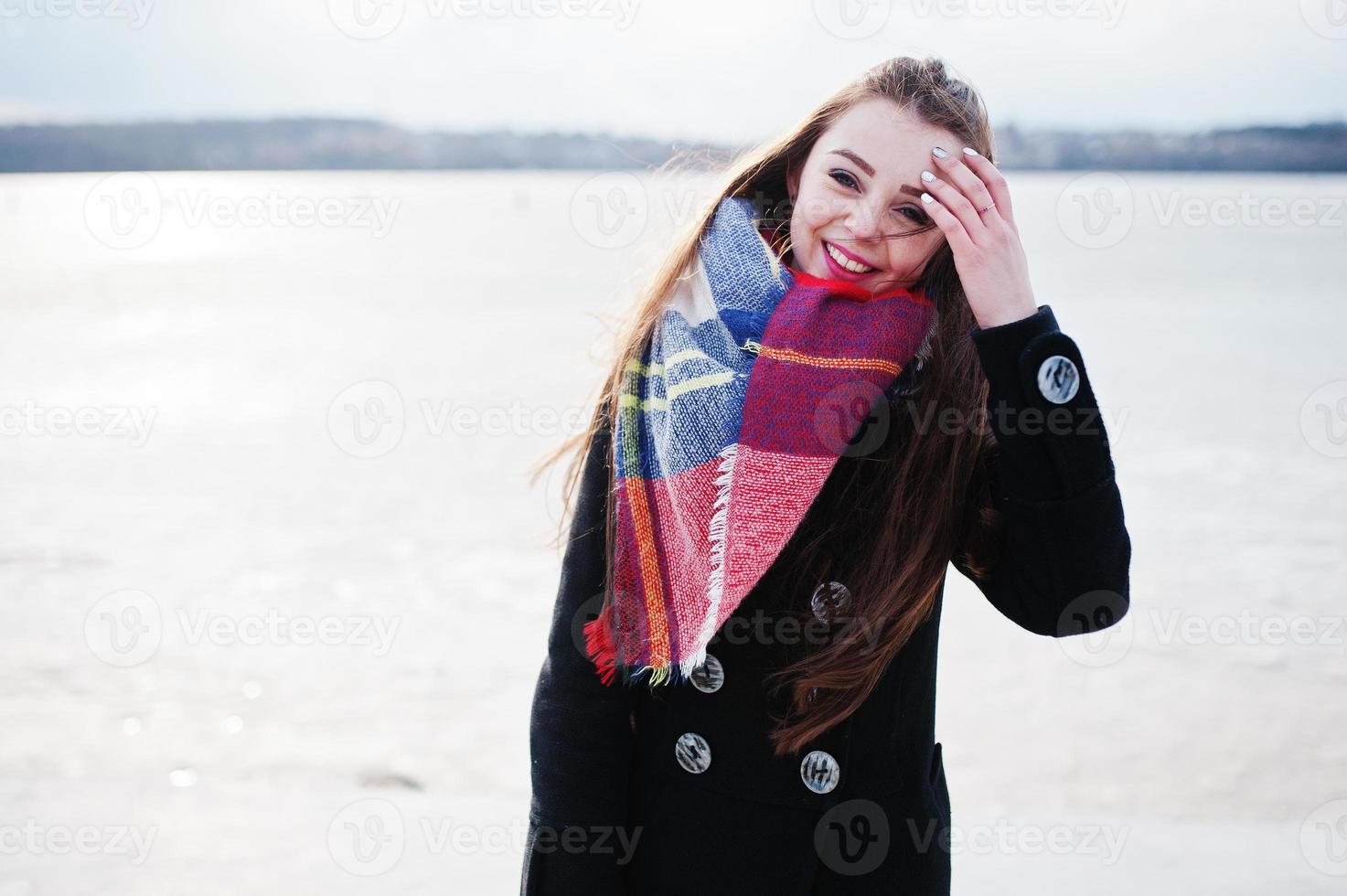 ragazza casual al cappotto nero, sciarpa e cappello contro il fiume ghiacciato in un clima invernale soleggiato. foto