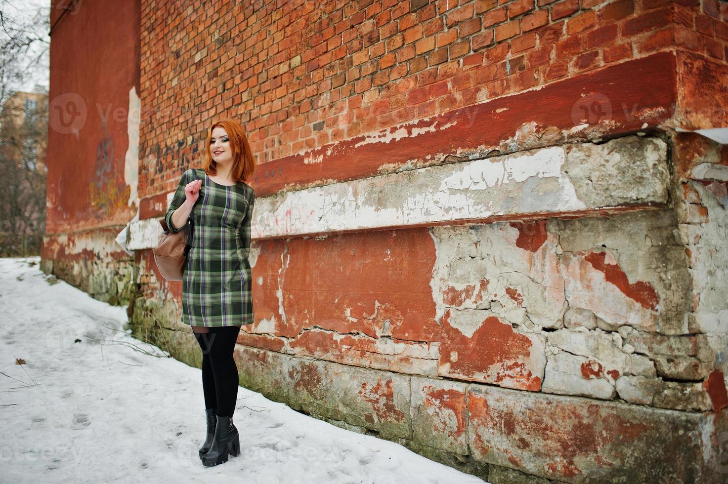 un ritratto all'aperto di una giovane bella ragazza con i capelli rossi che indossa un abito a scacchi con zaini girly in piedi sullo sfondo del muro di mattoni in una giornata invernale. foto
