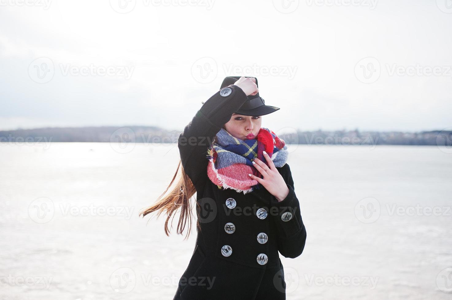 divertente ragazza casual al cappotto nero, sciarpa e cappello contro il fiume ghiacciato in un clima invernale soleggiato. foto