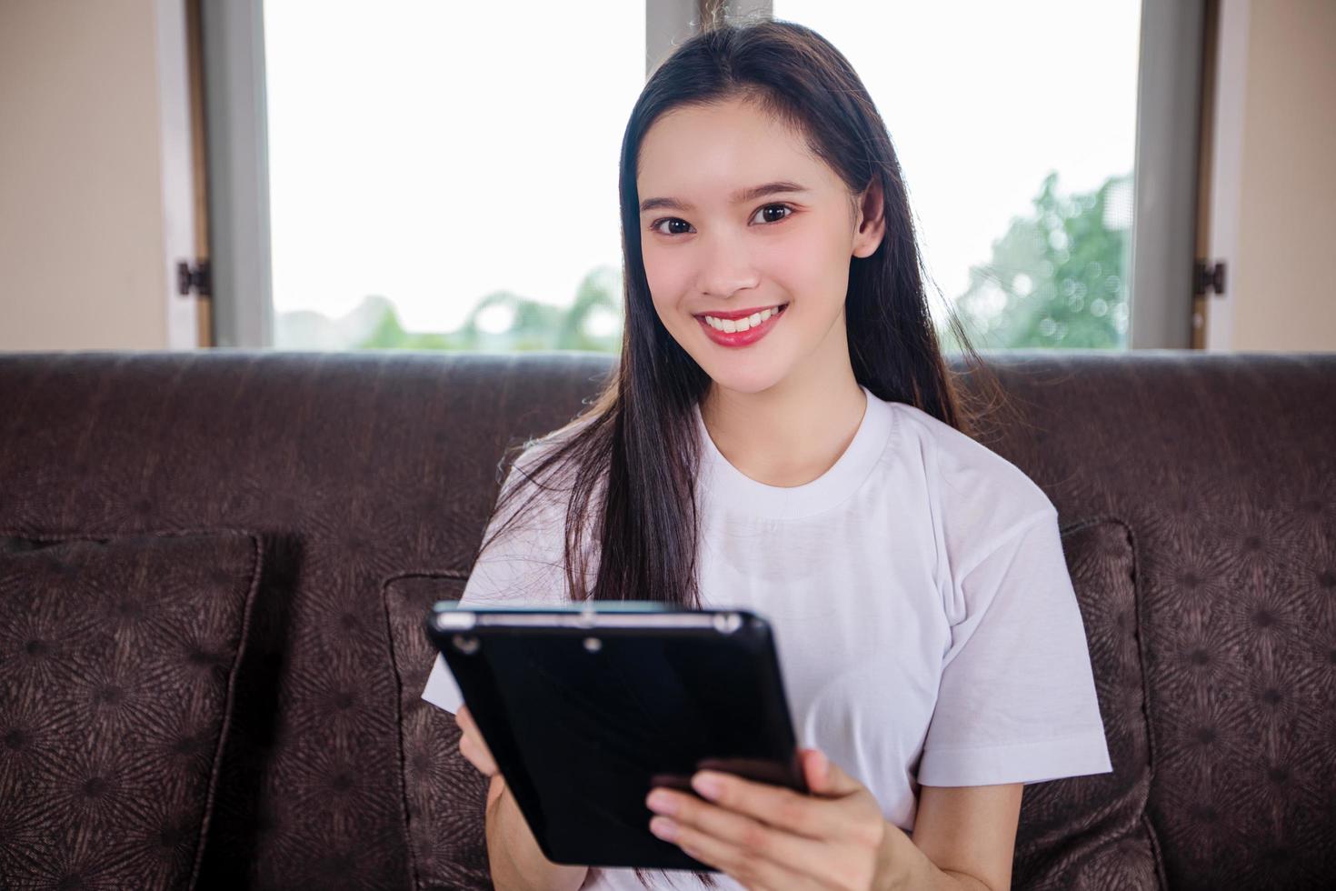 asiatico giovane donna felice allegro carino bello sta giocando digital pad tablet computer e seduto sul divano a casa, sorridente signora utilizzando app lettura libro elettronico rilassarsi in soggiorno foto