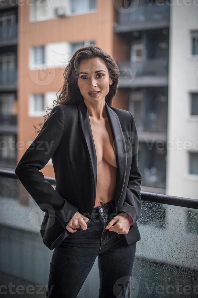 splendida donna bruna glamour con giacca nera in posa sul balcone moderno con splendida vista sulla città. ritratto di una donna alla moda alla moda con gambe lunghe, jeans neri che indossano in balcone foto