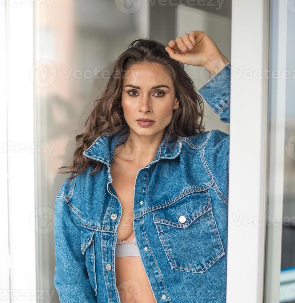 splendida donna bruna glamour con giacca blu in posa sul balcone moderno con splendida vista sulla città. ritratto di una donna alla moda alla moda con capelli lunghi, jeans neri che indossa in balcone foto