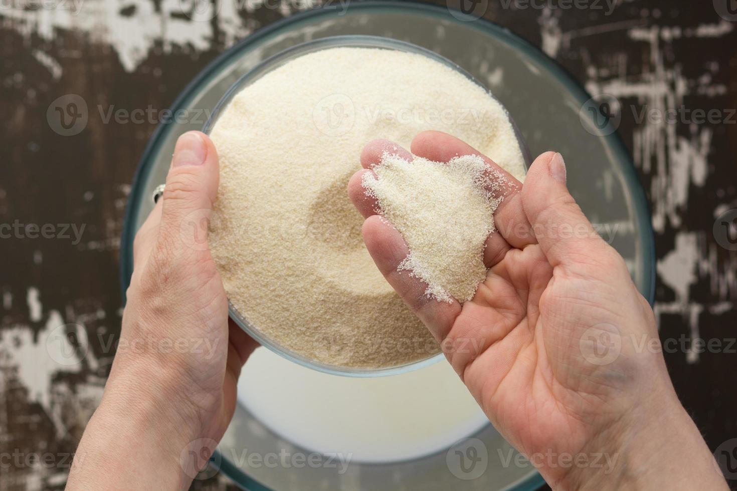 vista dall'alto della mano di womah che mette la semola nella ciotola con il latte come ingrediente per cuocere la casseruola mannik sullo sfondo marrone foto