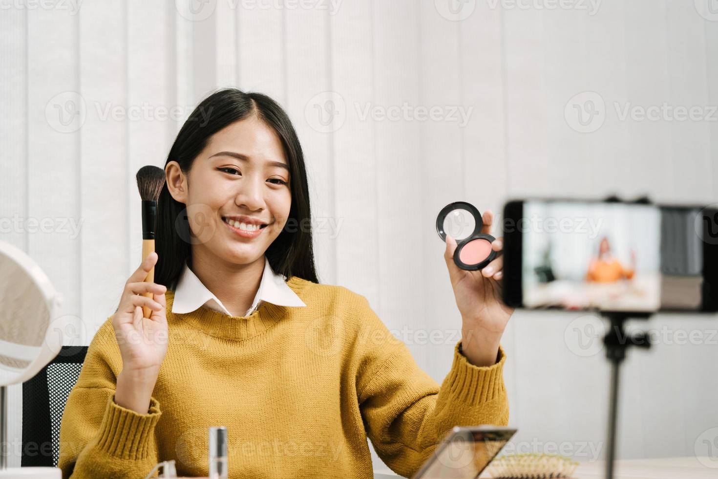 giovane donna asiatica che registra se stessa sui prodotti di bellezza realizzando un video per il suo blog sui cosmetici usando la fotocamera del telefono a casa. foto