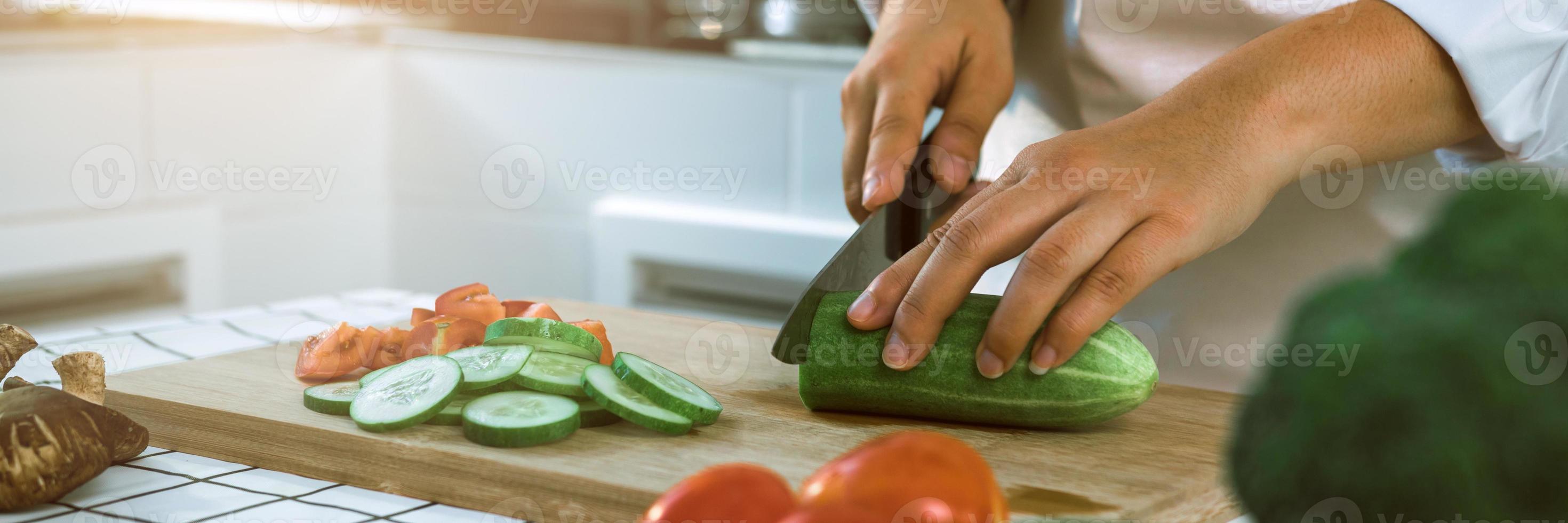 donna che usa coltello e mani che tagliano il cetriolo su tavola di legno nella stanza della cucina. foto