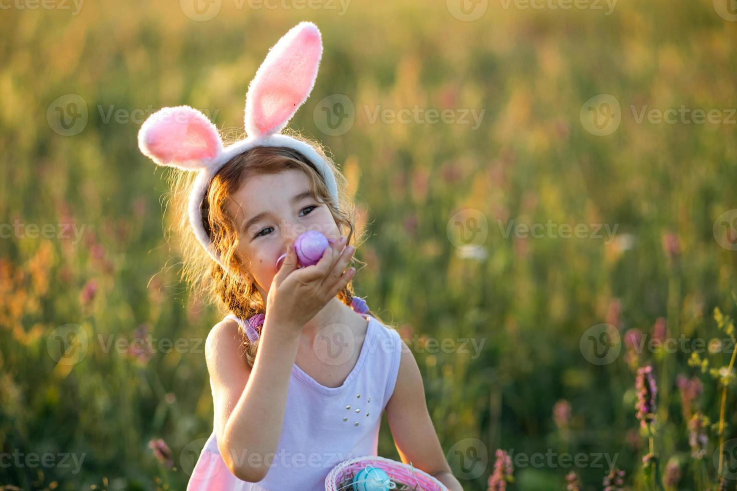 ragazza carina e divertente con uova di Pasqua dipinte in primavera in natura in un campo con luce solare dorata e fiori. vacanze di pasqua, coniglietto pasquale con le orecchie, uova colorate in un cestino. stile di vita foto