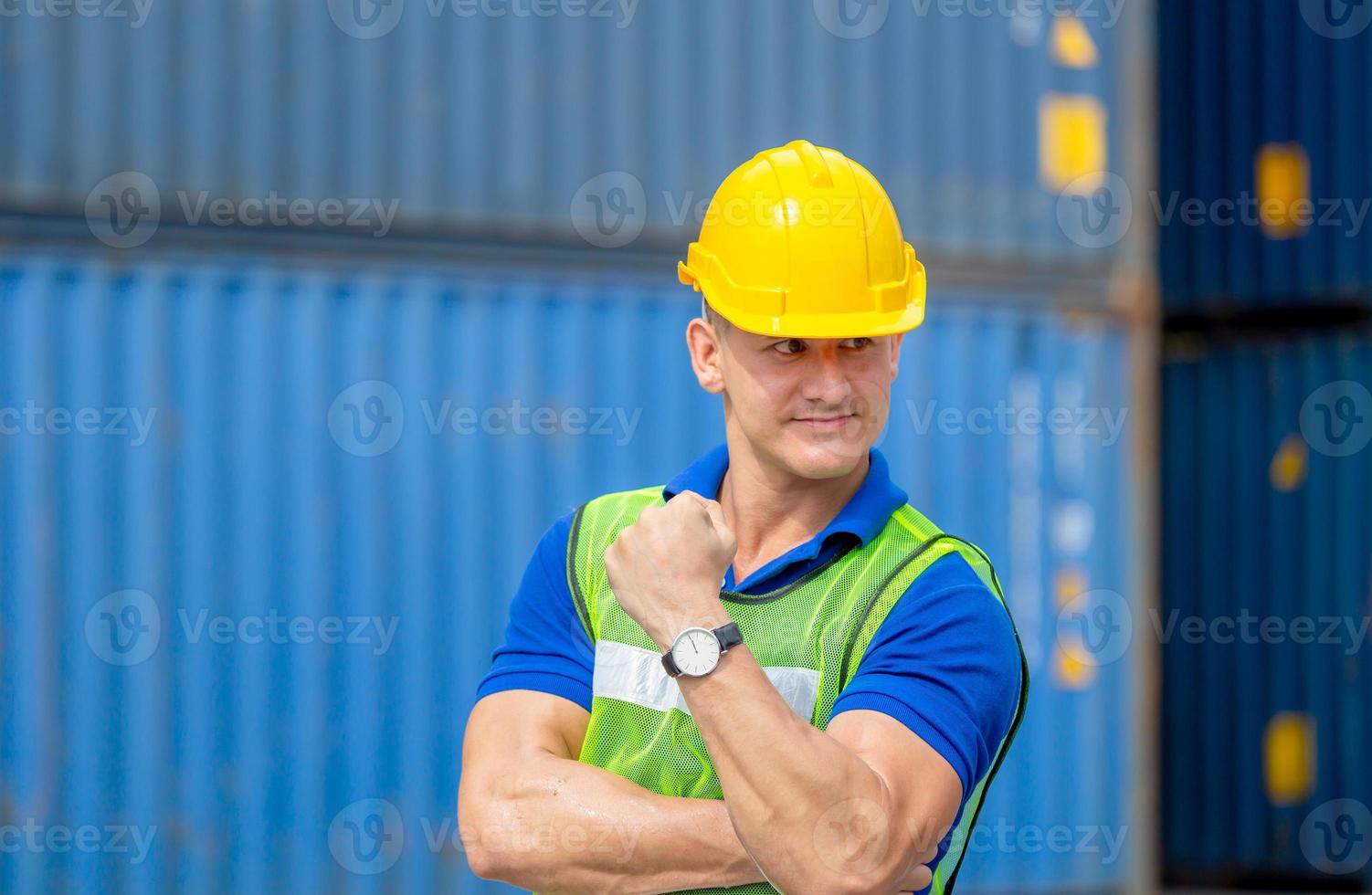 Felicità caposquadra lavoratore in elmetto protettivo mostra forti muscoli del braccio al carico di container in una giornata calda foto