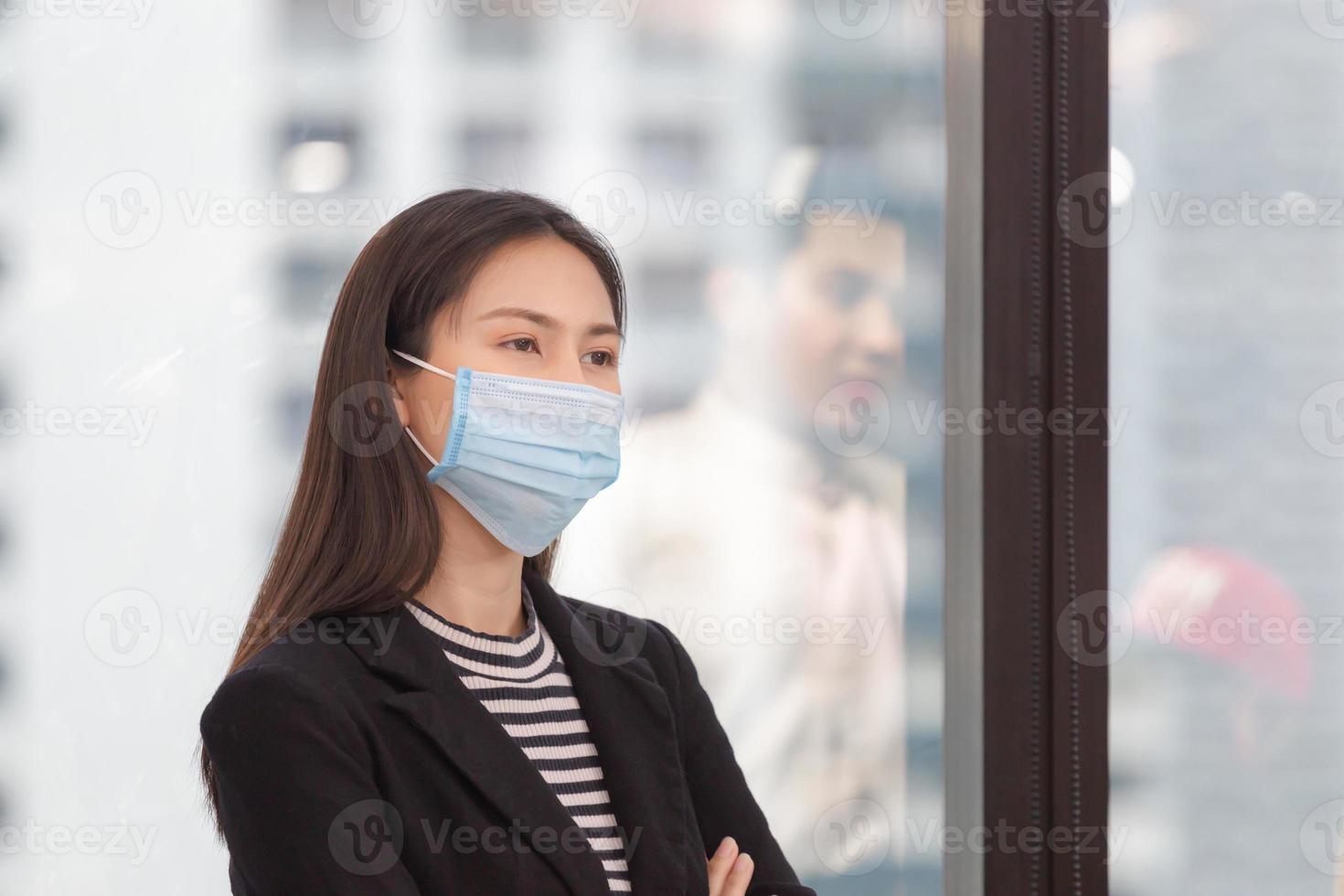 donna d'affari che indossa una maschera protettiva contro il coronavirus con le braccia incrociate in piedi in ufficio, nuovo concetto normale foto