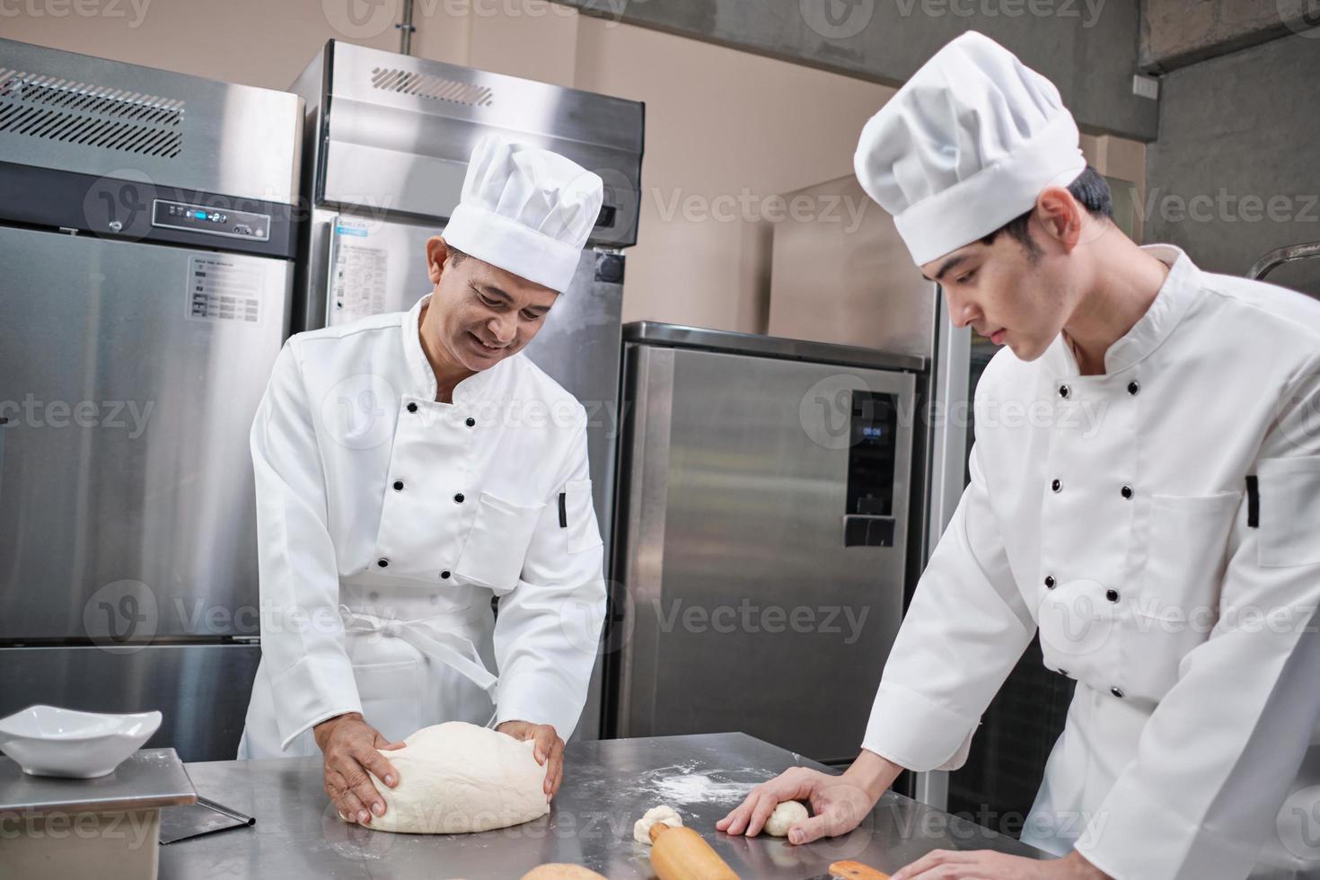 due cuochi professionisti asiatici maschi in uniformi bianche da cuoco e grembiuli stanno impastando pasta e uova, preparando pane e prodotti da forno freschi, cuocendo in forno nella cucina in acciaio inossidabile del ristorante. foto