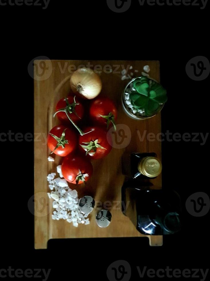 alcuni pomodori rossi freschi e una cipolla dorata adagiati su un tagliere di legno accanto a due bottiglie di olio e alcuni cristalli di sale bianco foto