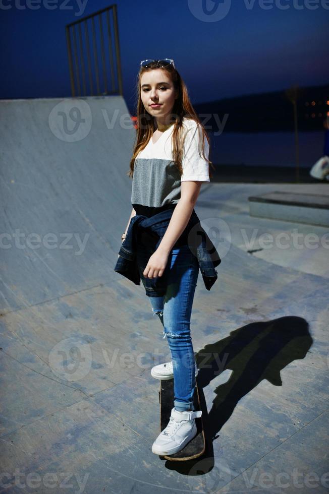 giovane ragazza urbana adolescente con skateboard, indossare occhiali, berretto e jeans strappati allo skate park la sera. foto