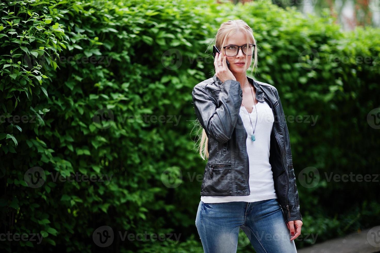 elegante donna bionda indossare jeans, occhiali e giacca di pelle con il telefono a portata di mano, contro i cespugli in strada. ritratto di modello urbano di moda. foto